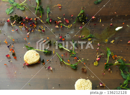 Leftover food scattered on a wooden table, featuring dirty dishes, remnants of a meal, and colorful edible flowers, creating a messy yet artistic culinary scene 136212128