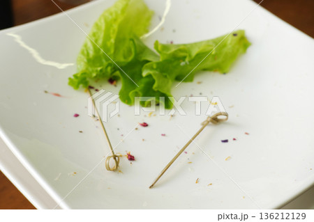 Leftover food remnants on a white plate, featuring wilted lettuce leaves and wooden skewers, highlighting food waste disposal and the impact of dirty dishes on the environment Leftover food remnants on a white plate, featuring wilted lettuce leaves and wooden skewers, highlighting food waste disposal and the impact of dirty dishes on the environment 136212129