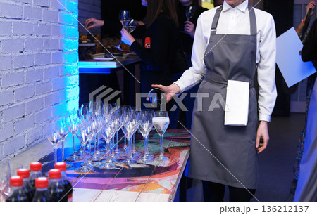 Professional catering service staff member in gray apron pouring drinks into elegant glasses at a stylish event, showcasing hospitality and attention to detail with copy space Professional catering service staff member in gray apron pouring drinks into elegant glasses at a stylish event, showcasing hospitality and attention to detail with copy space 136212137
