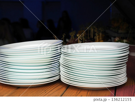 Stacked white plates with subtle green rims are arranged neatly on a wooden table, showcasing elegant tableware design and preparation for dining experience Stacked white plates with subtle green rims are arranged neatly on a wooden table, showcasing elegant tableware design and preparation for dining experience 136212155