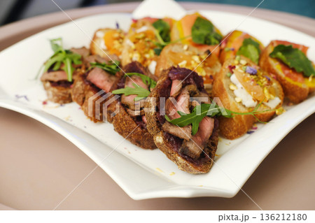 Gourmet platter featuring assorted canapes with grilled meat, colorful toppings, and fresh herbs, beautifully arranged on a white serving dish for an elegant dining experience Gourmet platter featuring assorted canapes with grilled meat, colorful toppings, and fresh herbs, beautifully arranged on a white serving dish for an elegant dining experience 136212180