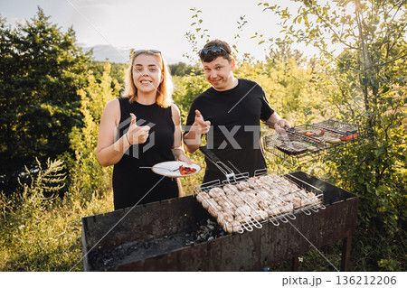 Couple giving thumbs up while grilling meat on outdoor barbecue at sunset 136212206