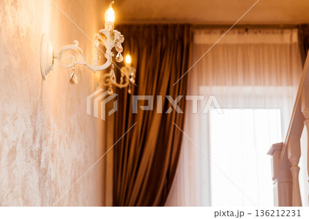 Elegant wall sconce with warm light illuminates a beautifully decorated interior space featuring rich drapery and soft textures, creating a cozy and inviting atmosphere Elegant wall sconce with warm light illuminates a beautifully decorated interior space featuring rich drapery and soft textures, creating a cozy and inviting atmosphere 136212231