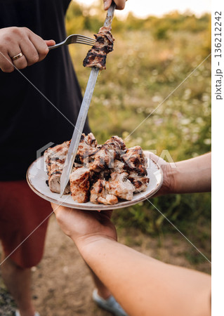 Serving grilled pork shashlik from skewer onto plate at outdoor barbecue 136212242