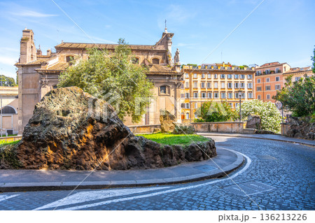 Visitors walk along Via di Monte Tarpeo in Rome, admiring the ancient architecture and natural scenery. The sunlight shines on a large rock and lush greenery near the street. 136213226