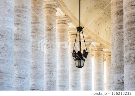 Visitors walk through the colonnade at St Peters Square in Vatican. The tall columns create a grand space under the ceiling. A light fixture hangs down, adding to the design of the area. 136213232