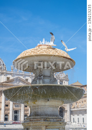 Water flows from the Bernini Fountain in St Peters Square. Birds stand on the fountain's edge. Visitors explore the square under a clear blue sky. The architectural details surround the area. 136213235
