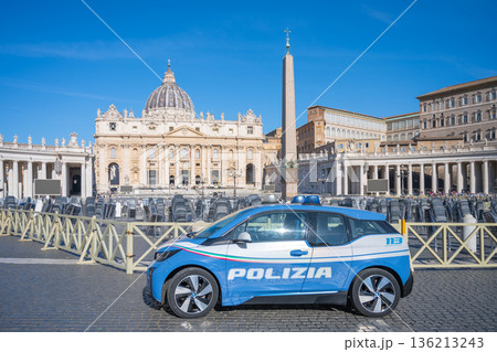 A police car is parked in St Peters square in Vatican city. Visitors and security barriers are present. The sun is shining on the iconic buildings in the background. A police car is parked in St Peters square in Vatican city. Visitors and security barriers are present. The sun is shining on the iconic buildings in the background. 136213243