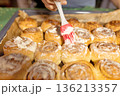Child's hand spreads frosting on homemade cinnamon buns made from yeast dough on gray baking sheet lined with parchment paper using red silicone brush, life style 136213357