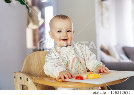 Smiling happy cute baby eating himself on high chair baby led weaning or blw. Infant self-feeding solid food fine motor development. Baby's feeding, learning eating food by himself and making mess 136213683