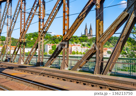 The Vysehrad Railway Bridge shows train tracks and metal structure. In the background, the Vltava River and buildings of Prague are visible under a clear sky. 136213750