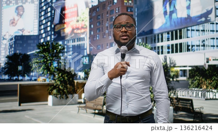 Adult male correspondent conducting public interview on city street, using confident dialogue to share news coverage. Demonstrating skill in storytelling in a metropolitan center. Camera A. 136216424