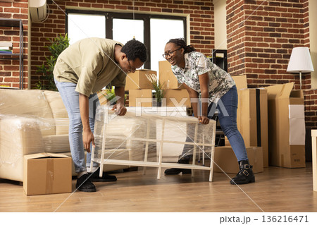 African american spouses happily carrying modern coffee table while relocating into new home. Excited married homeowners enjoying moving day, arranging furniture in fresh living room. 136216471