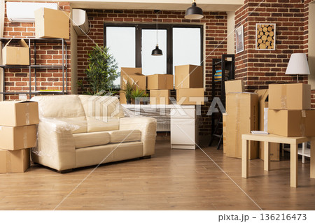 Vacant apartment living room with stacked cardboard boxes and furniture arranged near brick wall. Packed items awaiting movers as people prepare to move in or out of modern space. 136216473
