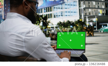 Green screen laptop with male typing emails and scrolling through online content while seated on a bench, his confident expression blending with the contemporary urban environment. Camera A. Green screen laptop with male typing emails and scrolling through online content while seated on a bench, his confident expression blending with the contemporary urban environment. Camera A. 136216486