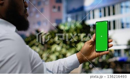 Vertical chroma key where african american man is holding his smartphone on a bench, sitting in a park near busy avenue. Modern lifestyle and calm attitude within city street activity. Camera B. Vertical chroma key where african american man is holding his smartphone on a bench, sitting in a park near busy avenue. Modern lifestyle and calm attitude within city street activity. Camera B. 136216493