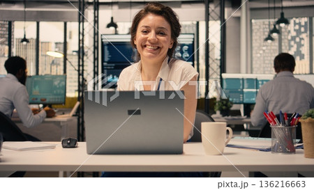 Portrait of businesswoman opening laptop seated at office desk, analyzing charts and graphs during performance review in a professional workspace. Corporate lifestyle with KPI metrics. 136216663