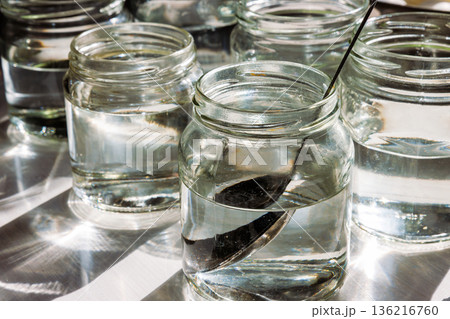 Close up of small glass jars filled with water in bright sunlight, with a spoon in one jar for stirring food coloring for easter egg dyeing 136216760