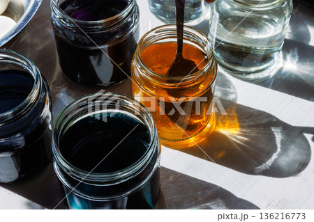 Top view of a glass jars with yellow and purple food dye dissolving in water, while someone is stirring the liquid it with a spoon 136216773