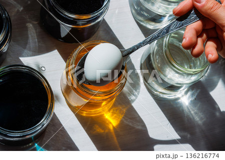 Top view of a white egg held on a spoon above a jar of yellow food dye, ready to dip, surrounded by clear water jars in bright sunlight. Top view of a white egg held on a spoon above a jar of yellow food dye, ready to dip, surrounded by clear water jars in bright sunlight. 136216774