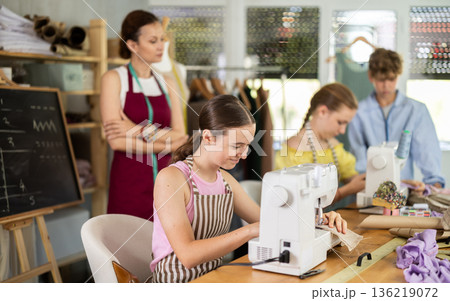 Teenage girl works on sewing machine while teacher and children learn how to cut fabric 136219072