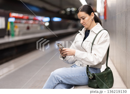 Girl with phone is sitting on metro platform station, scrolling though internet Girl with phone is sitting on metro platform station, scrolling though internet 136219130