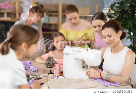 Woman teacher demonstrates how to sew on a sewing machine. Children gather around and watch Woman teacher demonstrates how to sew on a sewing machine. Children gather around and watch 136219189