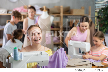 Teenage girl works on sewing machine while teacher and children learn how to cut fabric 136219376