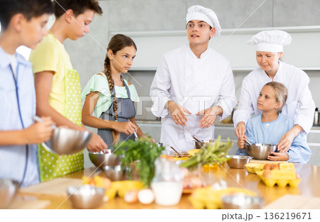 During cooking lesson, male cook tells children about culinary rules, woman assistant helps child 136219671