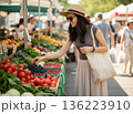Young woman in a straw hat, sunglasses and carrying a reusable eco-bag picking fresh grapes at a sunny outdoor farmers market. 136223910