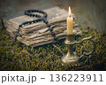 Old weathered book, a lit candle in a vintage brass holder and a black rosary with antique cross resting on green moss in a natural, soft-focus setting. 136223911