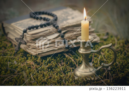 Old weathered book, a lit candle in a vintage brass holder and a black rosary with antique cross resting on green moss in a natural, soft-focus setting. 136223911