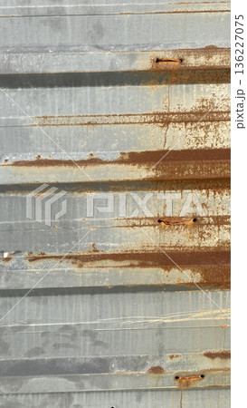 Close-up of a gray corrugated metal wall with brown rust lines and aged texture, ideal for industrial backgrounds.  136227075