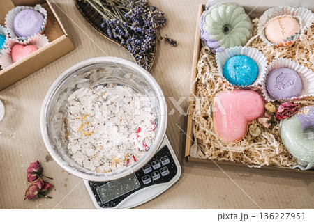 bowl containing dry bath bomb mixture sits on digital scale next to gift box of colorful heart and donut shaped soaps. Handmade bath bombs, bath salt diy, colorful self care gifts, bath art aesthetic. bowl containing dry bath bomb mixture sits on digital scale next to gift box of colorful heart and donut shaped soaps. Handmade bath bombs, bath salt diy, colorful self care gifts, bath art aesthetic. 136227951