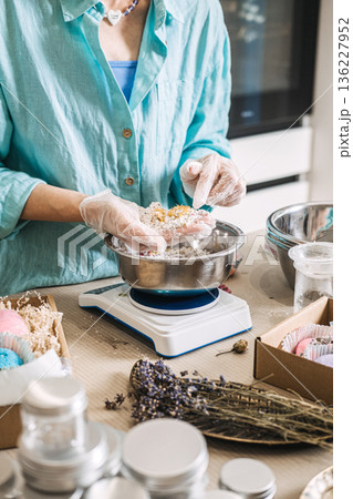 Hands hold large white bath bomb over metal bowl on digital scale surrounded by gift boxes. Small business scale, bulk bath bombs, production day vlog, handmade workshop. 136227952