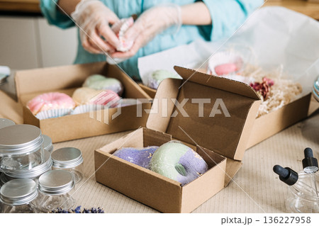 Person places colorful handmade soap products into cardboard boxes with paper filler and clear jars. Handmade bath bombs, personalized self-care gifts, bath salt mixology, aesthetic gift packaging. 136227958