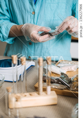 Hands in gloves pour dark minerals from small packet into glass test tube on digital scale. Waterless concentrate, anhydrous DIY, preservative-free bath, solid mineral skincare. 136227966