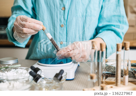 Gloved hands hold glass test tube filled with salt and botanical layers near digital scale. Anhydrous skincare, solid bath salts, waterless concentrate, botanical layering. 136227967