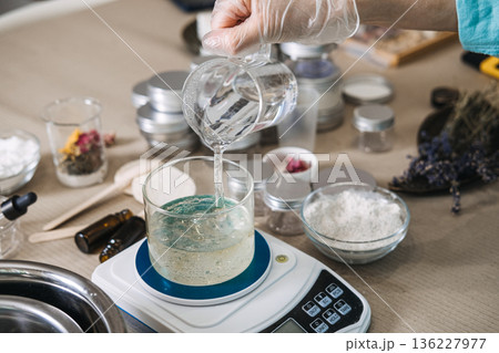Hand wearing glove pours clear liquid from beaker into glass jar on digital scale. micro-batch beauty, small business production, handcrafted moisturizer, artisanal beauty lab. 136227977