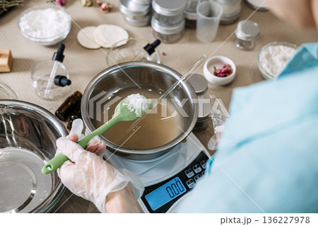 Person uses green spatula to hold white crystalline substance over metal bowl on digital scale. Hydration science aesthetic, pouring skincare liquid, water based cosmetic, refreshing beauty process. 136227978