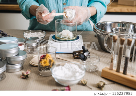 Hands in gloves scoop solid butter from jar into glass beaker on digital scale. raw beauty ingredients, skincare transparency trend, clean beauty formulation, botanical cosmetic actives. 136227983
