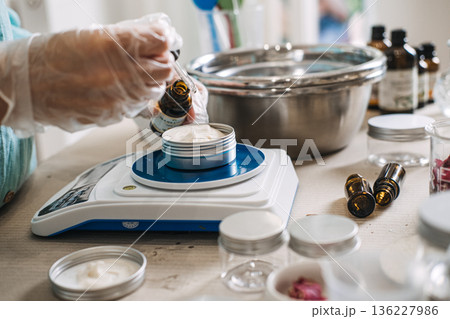 Person uses glass dropper to add oil to white cream in metal jar on digital scale. Lab-style beauty aesthetic, precise cosmetic formulation, diy apothecary lab, scientific skincare diy. 136227986