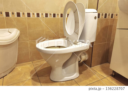 White toilet with open lid in a tiled bathroom interior, showing everyday sanitation, hygiene, plumbing fixture, and home restroom environment with neutral colors and practical design. White toilet with open lid in a tiled bathroom interior, showing everyday sanitation, hygiene, plumbing fixture, and home restroom environment with neutral colors and practical design. 136228707