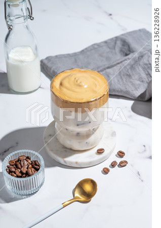 Dalgona coffee on a marble background with bottle, coffee beans, spoon and morning shadow. 136228926