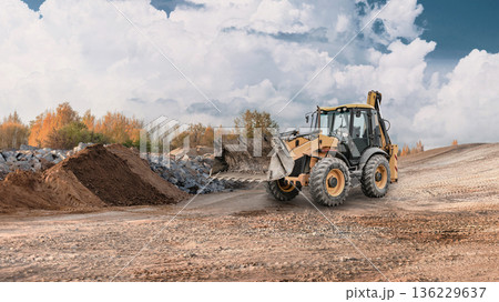 A wheel loader works at a construction site, moving soil and rock. The area is open, surrounded by piles of earth under a clear sky A wheel loader works at a construction site, moving soil and rock. The area is open, surrounded by piles of earth under a clear sky 136229637