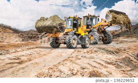 Two loaders carry sand and earth in buckets across a vast area of earthen banks, with clouds visible overhead 136229652
