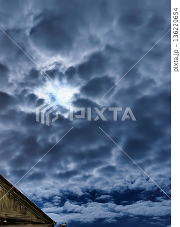 Puffy clouds fill the sky with a small area of sunlight breaking through. The scene depicts a late afternoon with a building in the foreground 136229654