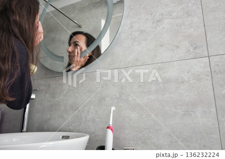 An attractive white Caucasian young woman looking in the bathroom mirror touching her wrinkles around her eyes - empty wall for copy space text An attractive white Caucasian young woman looking in the bathroom mirror touching her wrinkles around her eyes - empty wall for copy space text 136232224