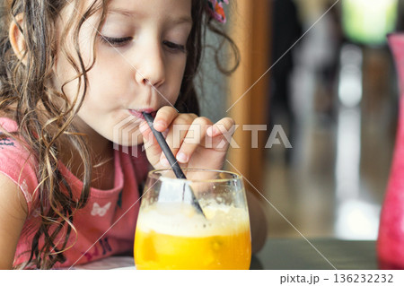 Close-up portrait of a cute little white Caucasian girl drinking a glass of fresh orange juice with a straw 136232232
