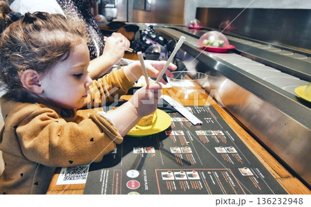 Cute little girl using chopsticks to eat sushi at a restaurant with self-service conveyor belt 136232948